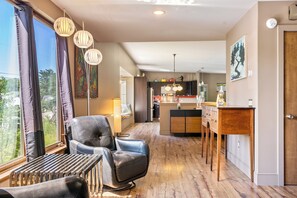 A modern living room with a leather sofa, a dining area with a wooden table, and a kitchen in the background. - The Crag: On the Acadia National Park Border - Deck, Fire Pit, Steps to Trails (Mount Desert)