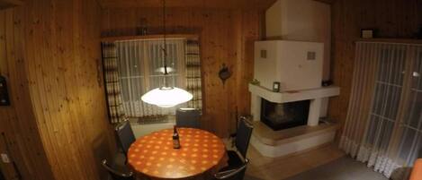 A wooden interior with a round dining table, chairs, a hanging light fixture, and a fireplace.