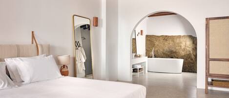 Un dormitorio moderno con una cama grande, cabecera de madera, una mesita de noche, un espejo y un arco que da a un baño con bañera.
