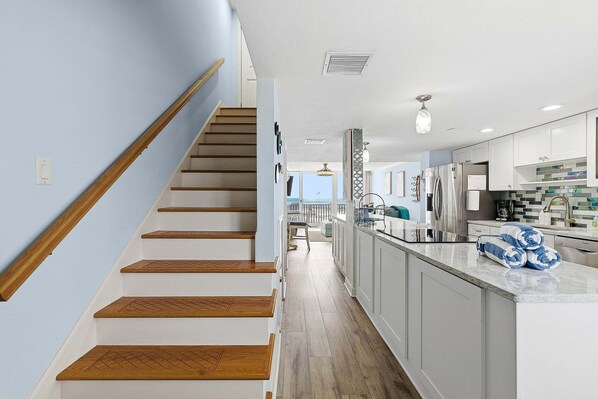 A modern kitchen with white cabinets, a wooden staircase, and a view to the living area. - Unobstructed Oceanfront Views | 2 Balconies | Quiet Coastal Escape (New Smyrna Beach)