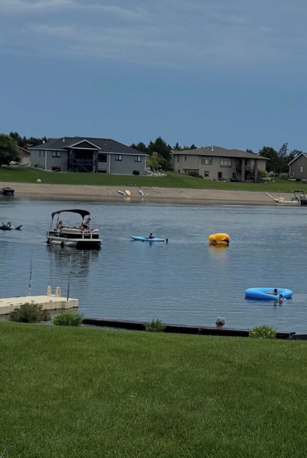 Un lago tranquilo con barcos, una orilla cubierta de hierba y casas al fondo.