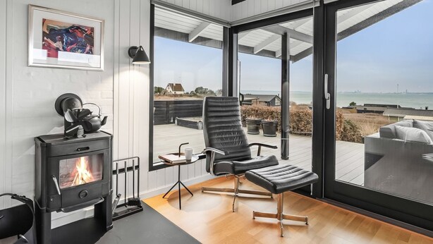 Een modern interieur met een houtkachel, een zwarte lederen fauteuil en een grote glazen deur die leidt naar een terras met uitzicht op zee.