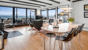 Een ruime kamer met een grote eettafel, zithoek, televisie en grote ramen met uitzicht op zee.