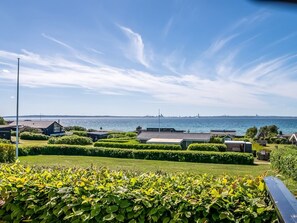 Ett bostadsområde vid kusten med välklippta häckar, fri utsikt över havet och en avlägsen stadsbild i horisonten.