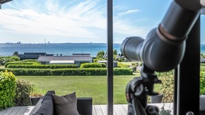 Een overdekt buitengebied met uitzicht op zee, een telescoop en verzorgde hagen.