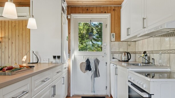Cocina compacta con armarios blancos, techo de madera, ventana con vistas a árboles y un lavamanos con hervidor.