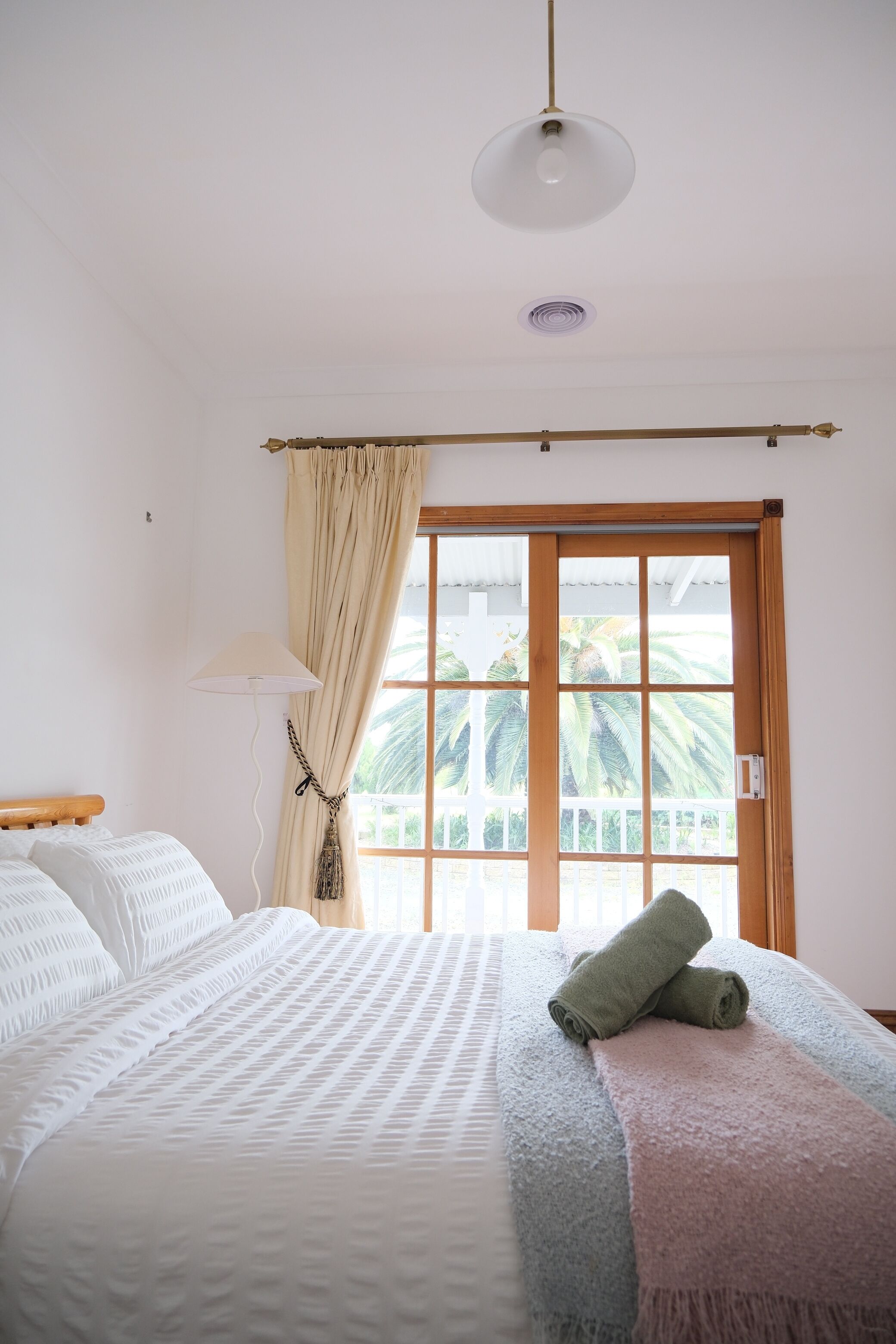 A bedroom with a bed, a window with curtains, a lamp, and a view of palm trees.
