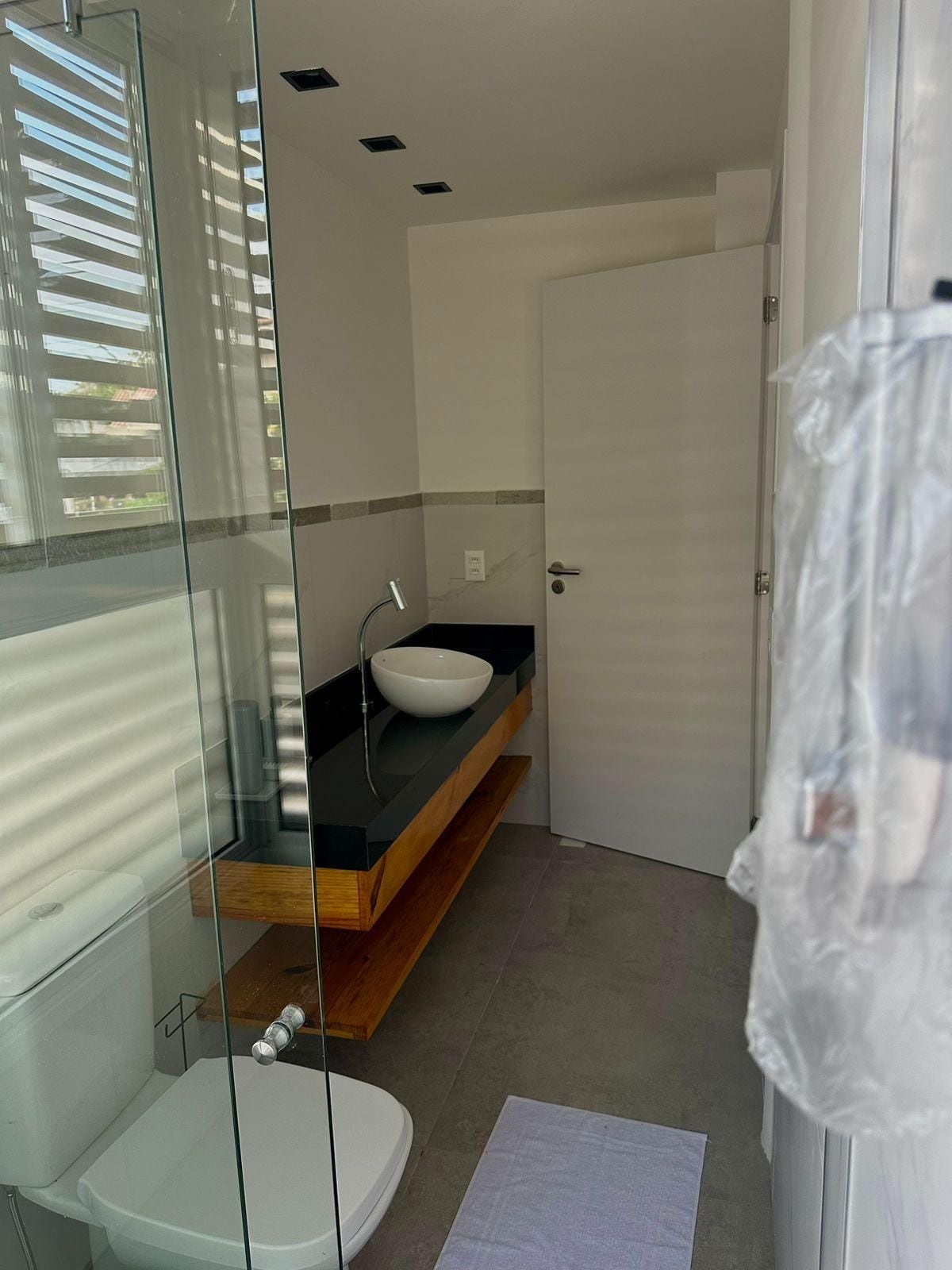 Un baño moderno con mampara de vidrio, inodoro blanco, lavamanos blanco y encimera negra, además de un estante de madera.