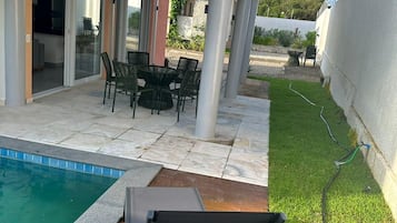 Un área junto a la piscina con una mesa y sillas, una pérgola y un césped.