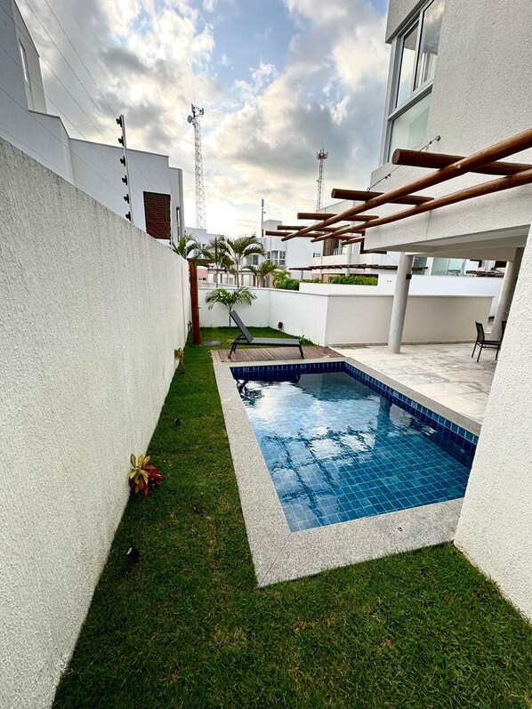 A residential outdoor area with a swimming pool, a grassy lawn, and a white wall.