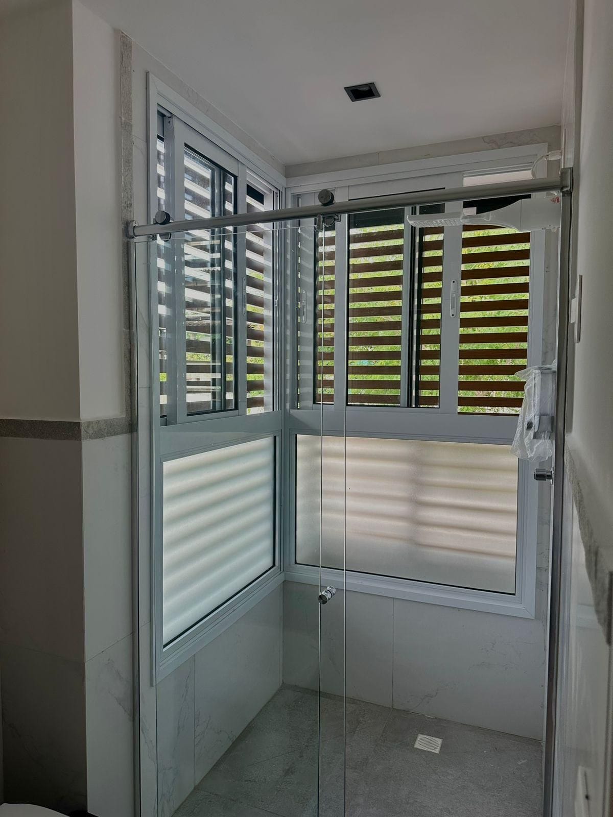 A corner bathroom with a glass shower enclosure, a window with blinds, and a tiled floor.