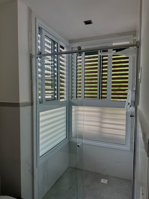 Un baño en esquina con cabina de ducha de vidrio, una ventana con persianas y suelo de baldosas.