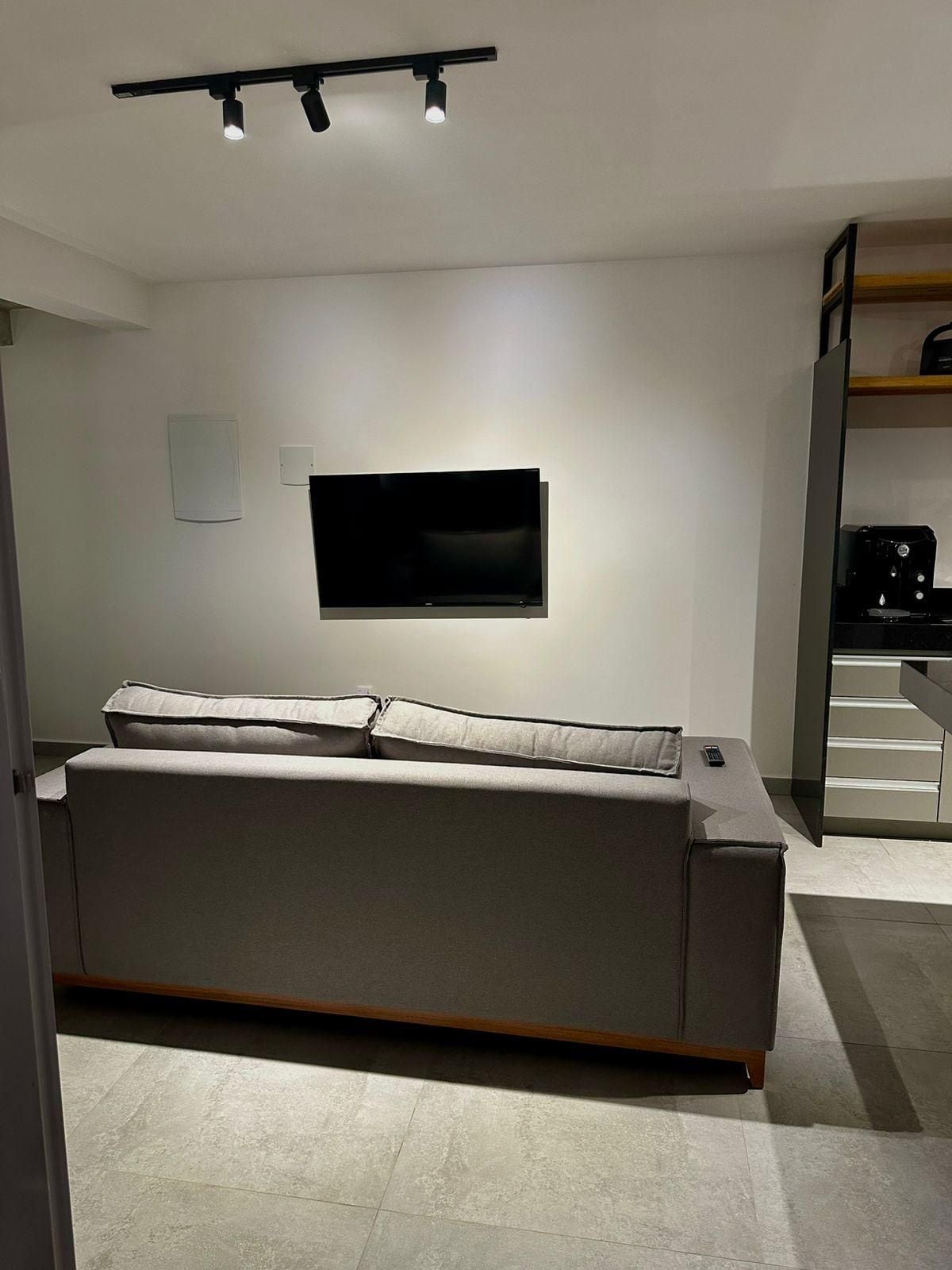 A modern living room with a sofa, a flat-screen TV mounted on the wall, and a coffee machine on a counter.