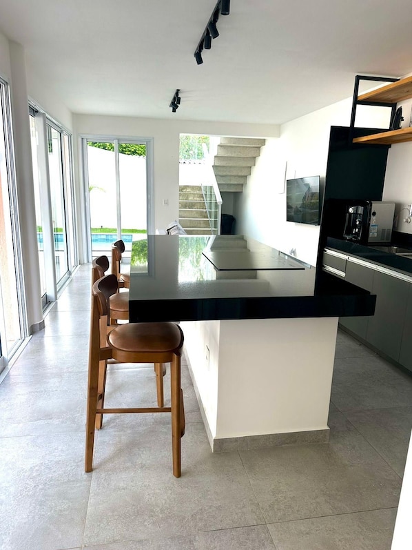 A modern kitchen with a large island, bar stools, a coffee machine, and a view of a staircase leading to a balcony. - Praia da Pipa - Mar e Descanso (Tibau do Sul)