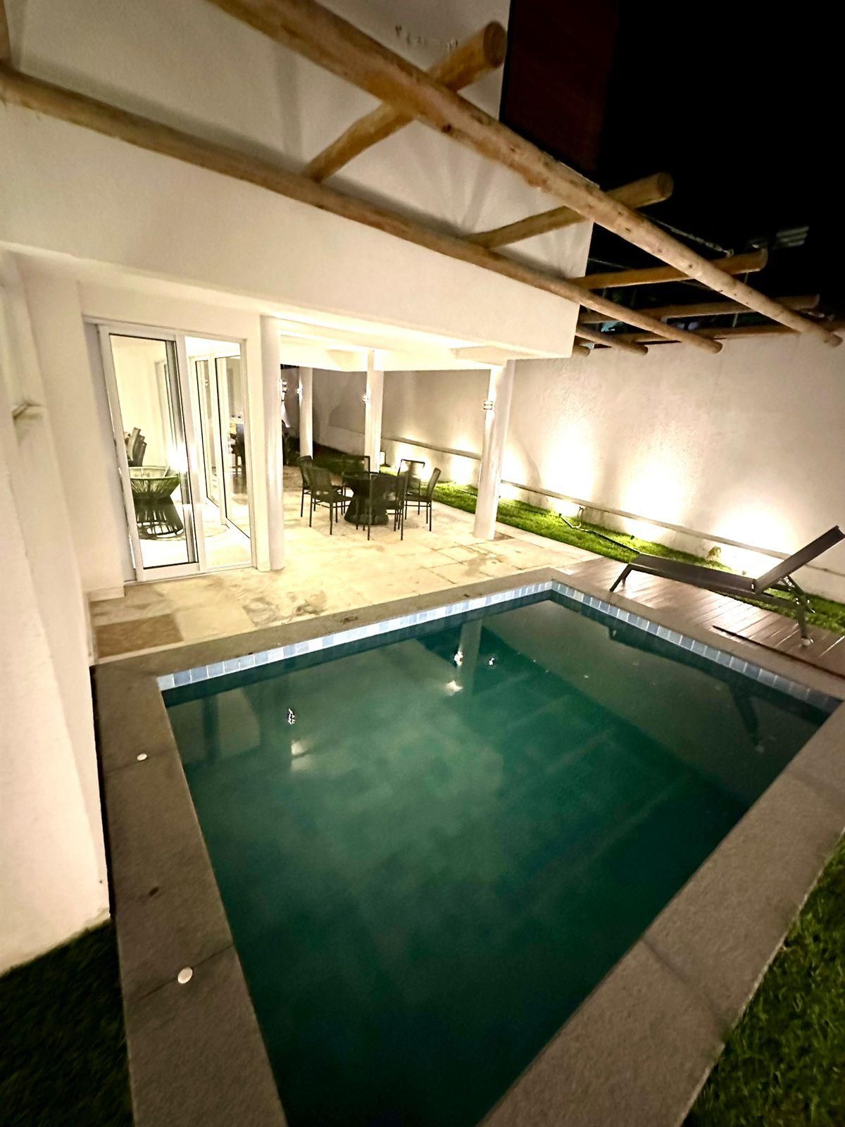 Una piscina con zona de patio, que incluye una mesa y sillas, y una sección cubierta con vista al interior.