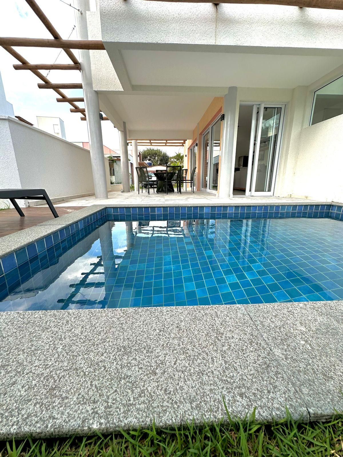 Uma piscina com borda azulejada, mesa e cadeiras sob uma pérgula, e um edifício com porta de vidro ao fundo.