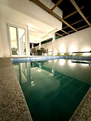 An indoor swimming pool with clear water, surrounded by a tiled deck and a seating area with chairs.