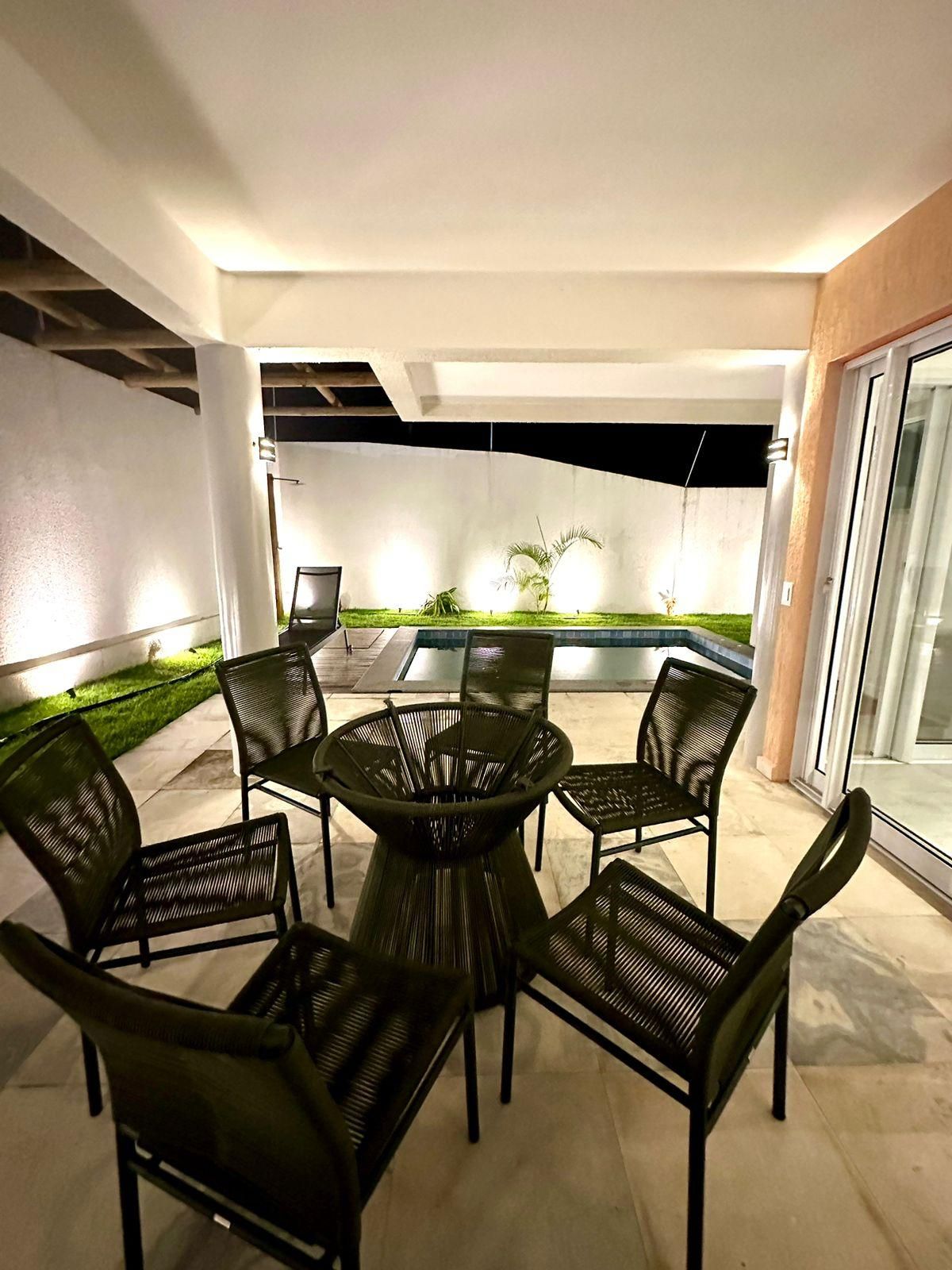 Uma área de pátio ao ar livre com mesa redonda e seis cadeiras, uma pequena piscina e uma área gramada com plantas.