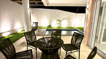 Uma área de pátio ao ar livre com mesa redonda e seis cadeiras, uma pequena piscina e uma área gramada com plantas.