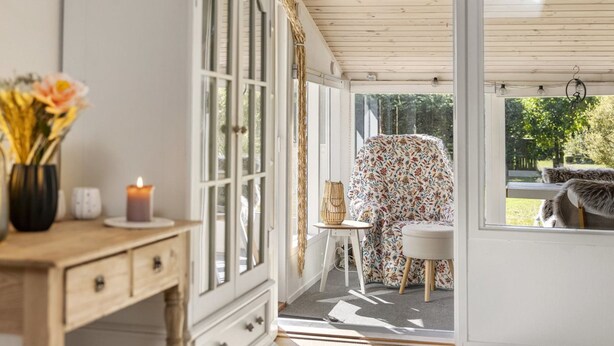 A room with a wooden desk, a cabinet with glass doors, a chair covered in floral fabric, and a view of the outdoors through large windows. - "Ansfried" - 900m from the sea by Interhome (Sæby)