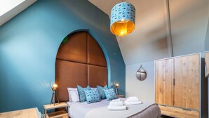 Quarto moderno com teto inclinado, cama grande com almofadas estampadas, abajures, guarda-roupa de madeira e luminária pendente.