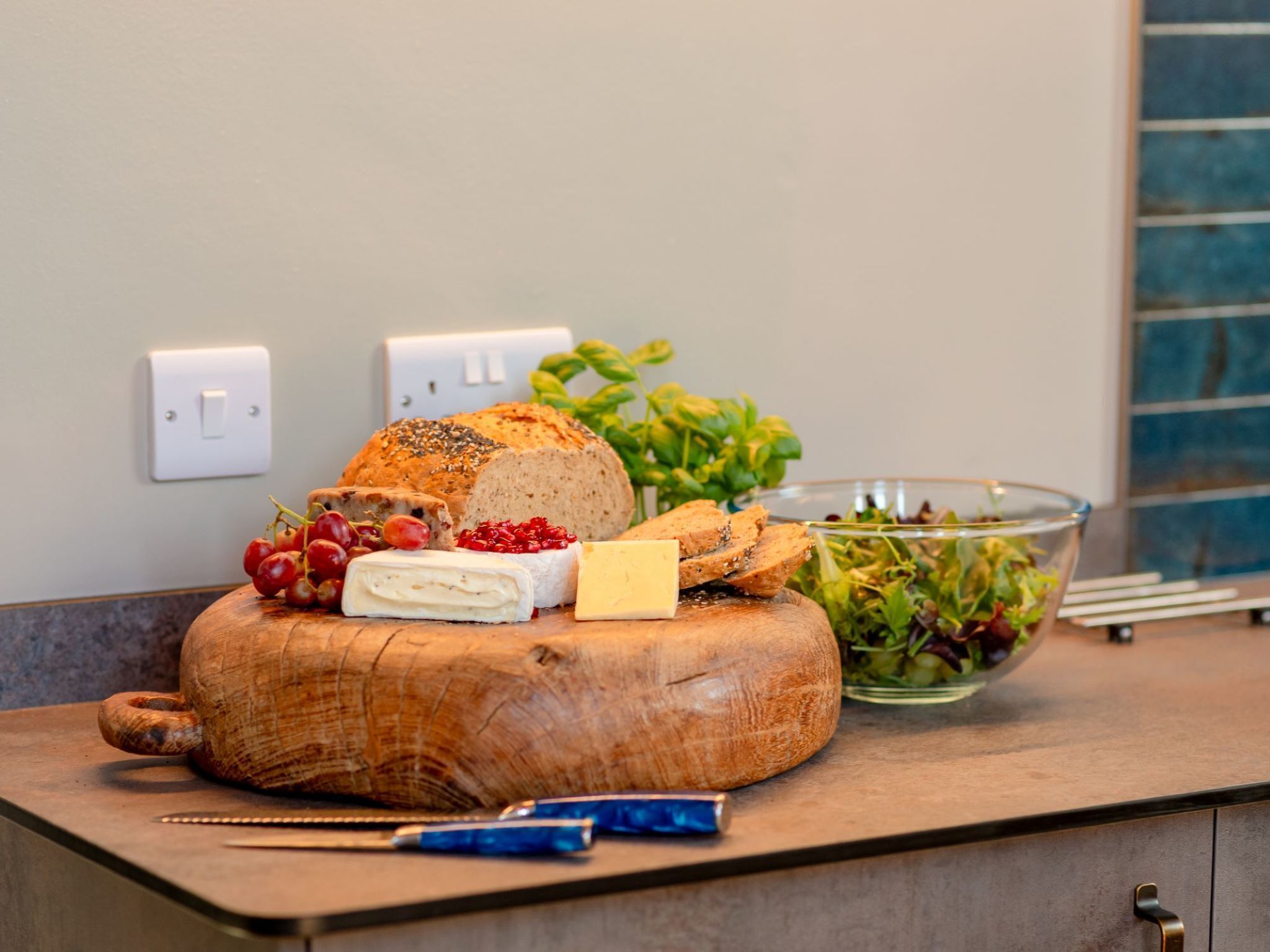 A wooden cutting board with cheese, bread, and grapes on a kitchen counter.