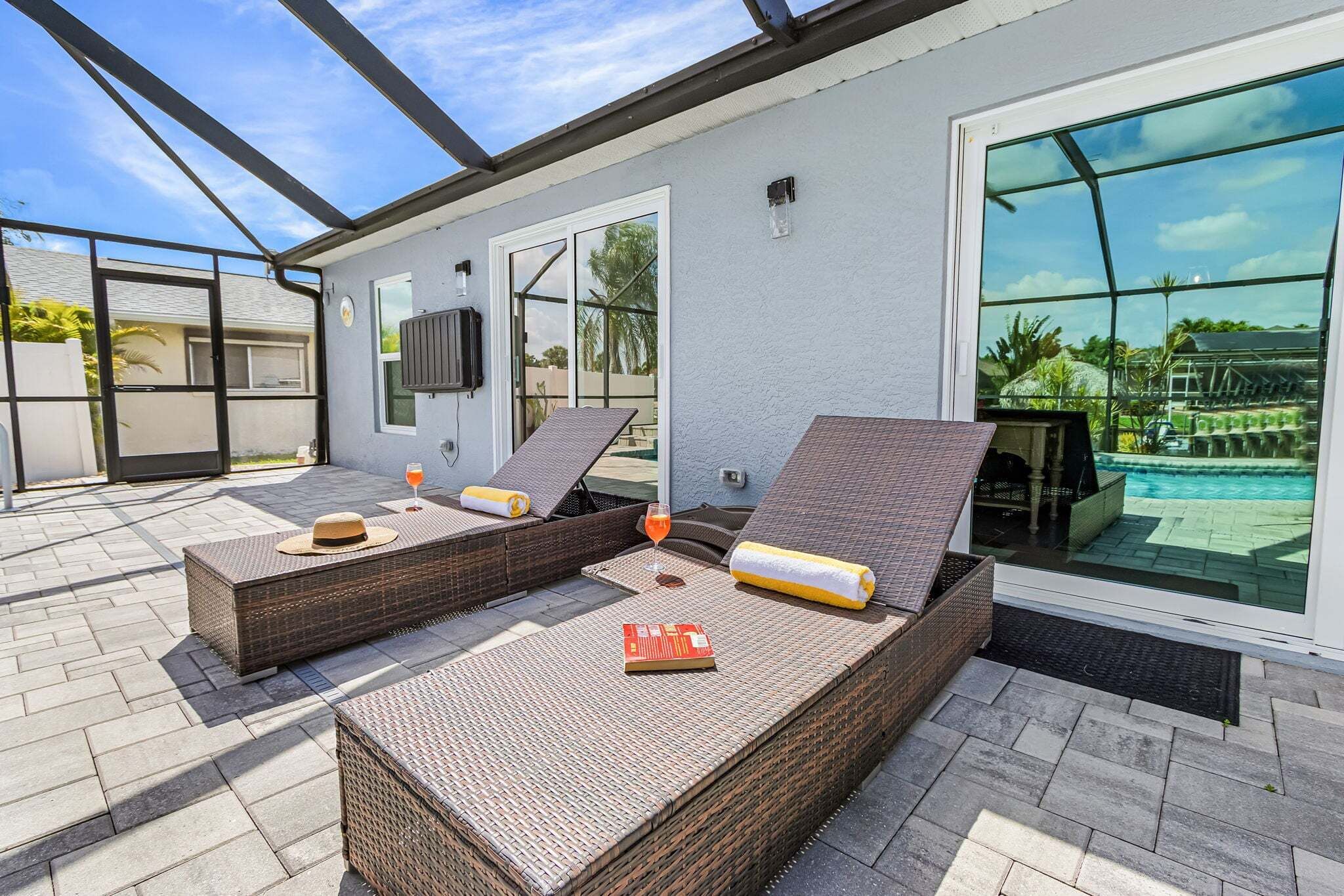 A sunlit outdoor lounge area with wicker furniture, a glass door leading to a pool, and a clear view of the surrounding landscape.