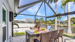 Un patio exterior con una parrilla, una mesa de comedor y sillas, con vista a un cuerpo de agua y palmeras.