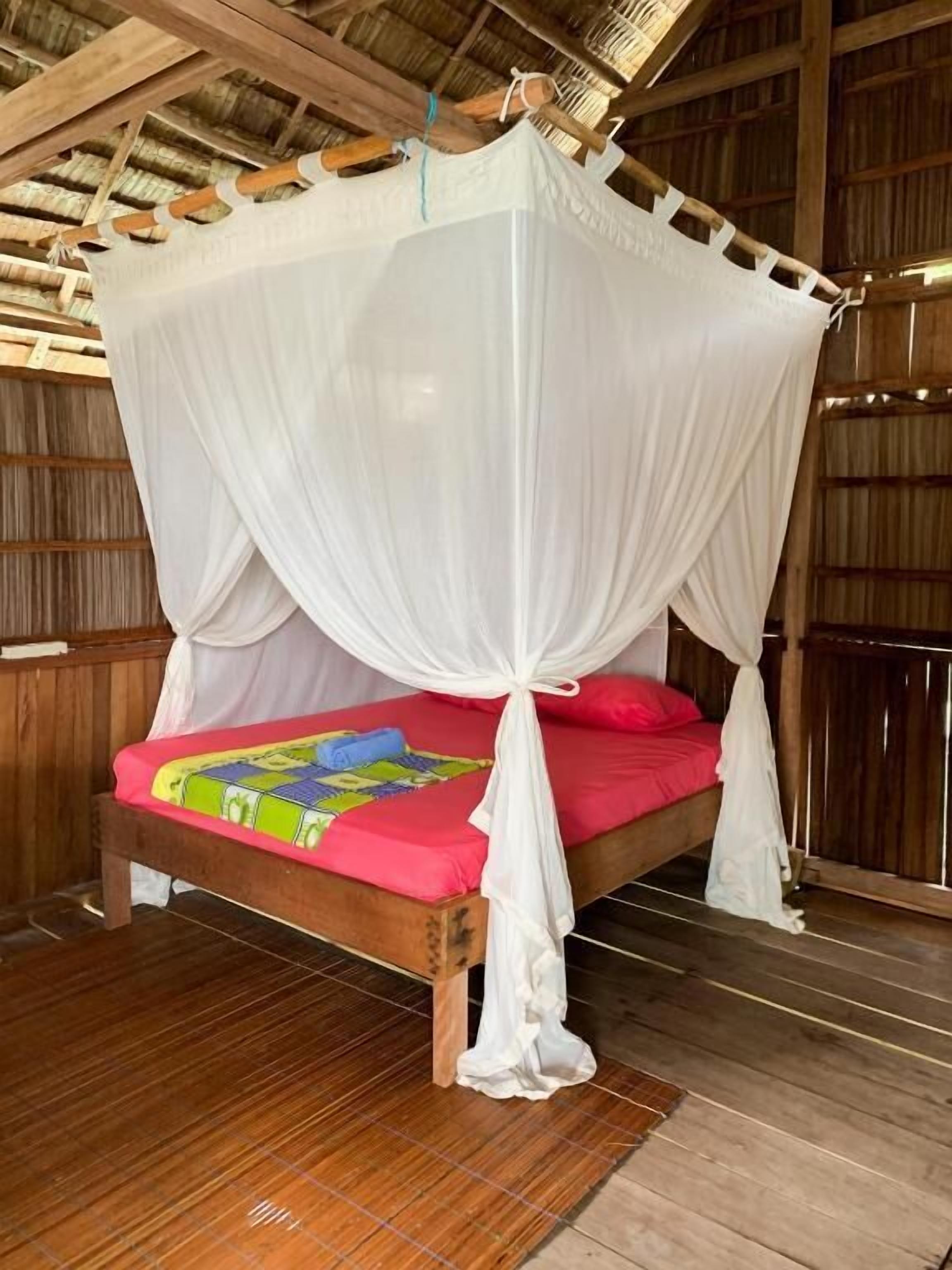 A bed with a white canopy, a wooden floor, and a thatched roof.