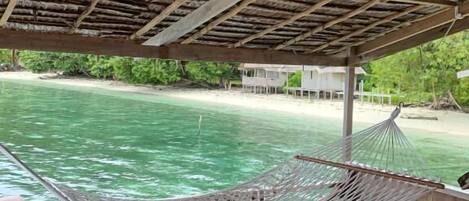 A hammock under a thatched roof, overlooking a beach.