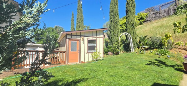 Une petite maison avec une porte et des fenêtres marron, entourée d’une pelouse bien entretenue et de grands arbres.