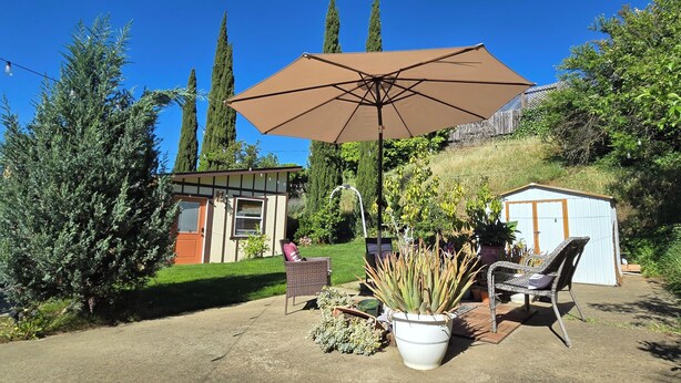 Un patio con una sombrilla grande, mobiliario de mimbre, plantas en macetas y un cobertizo pequeño.