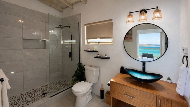 Een moderne badkamer met een glazen douche, toilet, wastafel met een blauwe kom en een spiegel met uitzicht op zee.