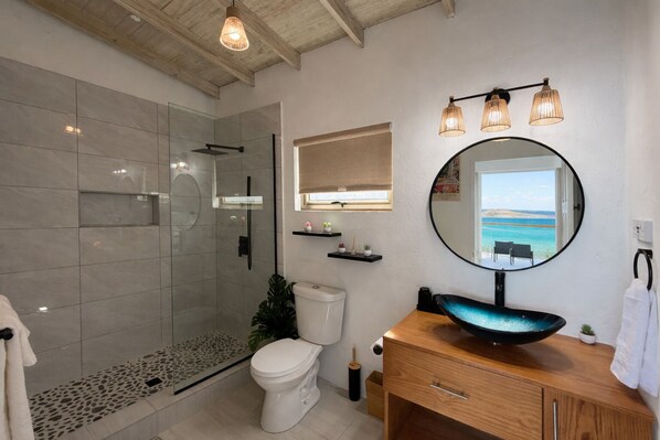 Une salle de bain moderne avec une douche en verre, des toilettes, un lavabo avec un receveur bleu et un miroir offrant une vue sur la mer.