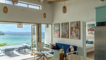 A room with a view of the sea, a dining area with a table and chairs, and a sofa with cushions.