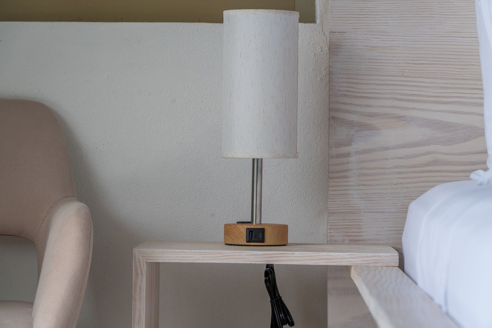 A bedside table with a lamp, a chair, and a bed in a hotel room.