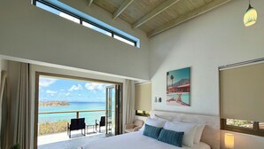 Een slaapkamer met een groot bed, een nachtkastje met een lamp, een schilderij en uitzicht op de zee door een schuifdeur.