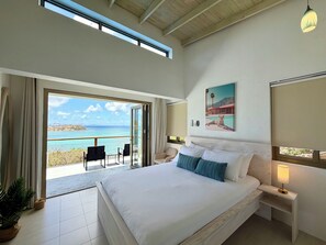 Une chambre à coucher avec un grand lit, une table de chevet avec une lampe, un tableau et une vue sur la mer, visible à travers une porte coulissante.