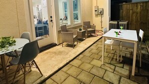 Een terras buiten met stenen vloer, een overdekte zithoek met rieten stoelen en een witte tafel.