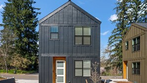 A two-story house with a dark exterior, a front door, and two windows with shutters. - Chiquita Bonita | Brand New, Stylish & Spotless, Pet Friendly, Walk to Park, Minutes from Downtown (Tigard)