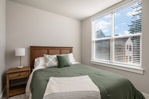 A bedroom with a bed, nightstand, lamp, and window with blinds. - Chiquita Bonita | Brand New, Stylish & Spotless, Pet Friendly, Walk to Park, Minutes from Downtown (Tigard)
