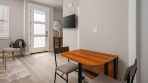 A modern room with a wooden dining table, two chairs, a TV mounted on the wall, and a door leading to another room. - Chiquita Bonita | Brand New, Stylish & Spotless, Pet Friendly, Walk to Park, Minutes from Downtown (Tigard)