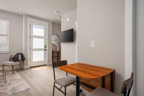 Quarto moderno com mesa de jantar de madeira, duas cadeiras, televisão na parede e porta para outro espaço.