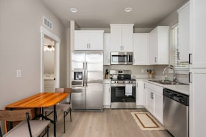 A modern kitchen with white cabinets, stainless steel appliances, a wooden dining table, and a small rug. - Chiquita Bonita | Brand New, Stylish & Spotless, Pet Friendly, Walk to Park, Minutes from Downtown (Tigard)