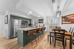 Eine moderne Küche mit einem Edelstahlkühlschrank, einer Mikrowelle und einer Kaffeebar. Es gibt eine Bar mit Hockern sowie einen Esstisch mit Stühlen.