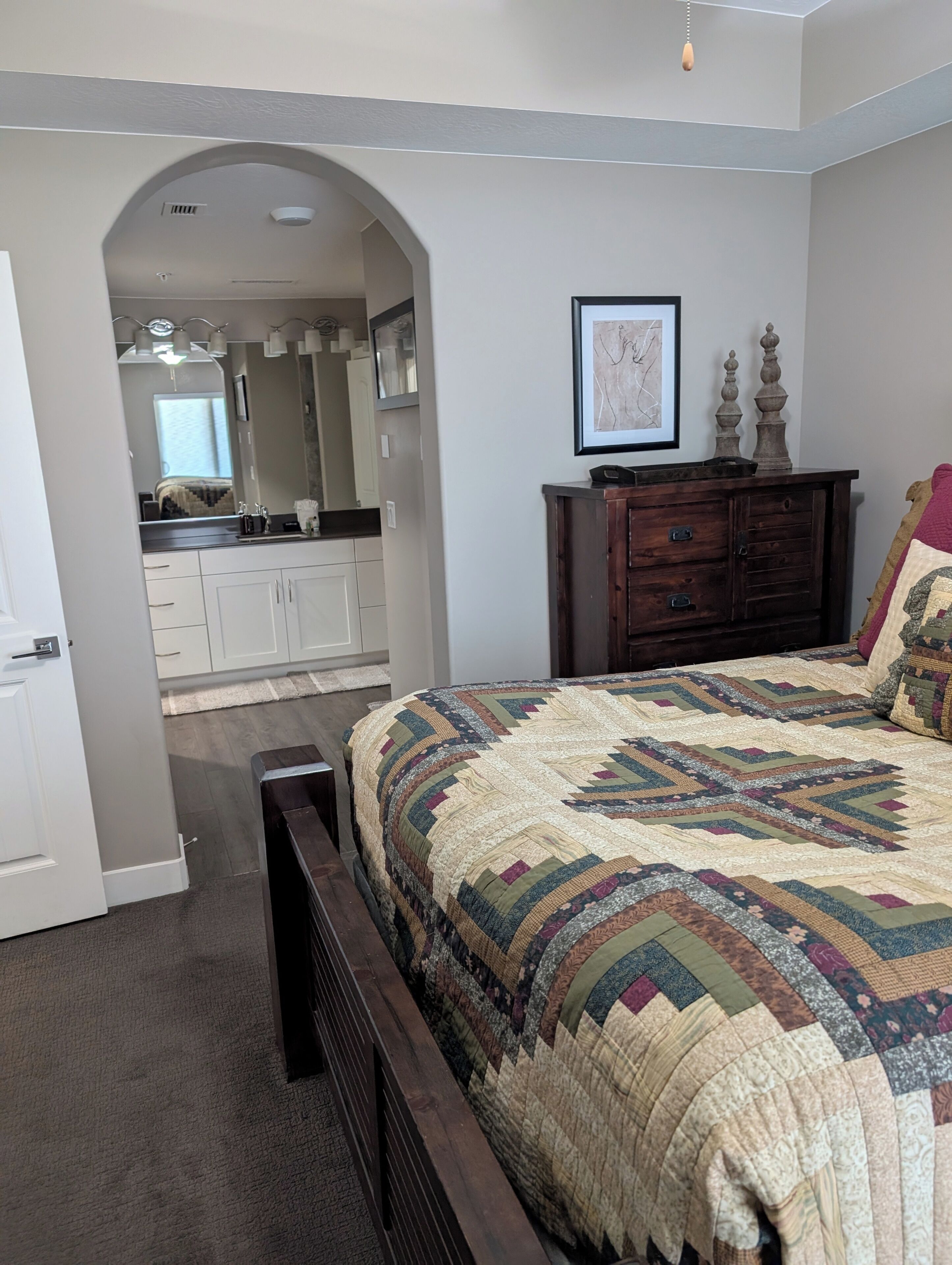 A bedroom with a large bed, a dresser, a mirror, and an arched doorway leading to a bathroom.