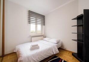 Una camera da letto con un letto, un termosifone, una finestra con tende e una scaffalatura nera, il pavimento è in legno.