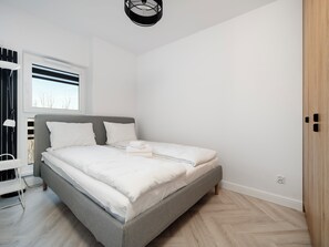 Une chambre moderne avec un lit gris, du linge de lit blanc, une fenêtre avec des stores, une table de chevet et un luminaire suspendu.