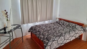 A bedroom with a bed, a small table with a vase and reed diffuser, a window with curtains, and a tiled floor.
