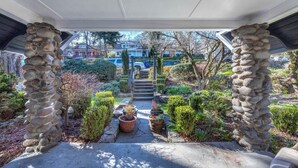 Arcada de pedra que dá para um jardim com plantas em vasos e vista para uma rua residencial.